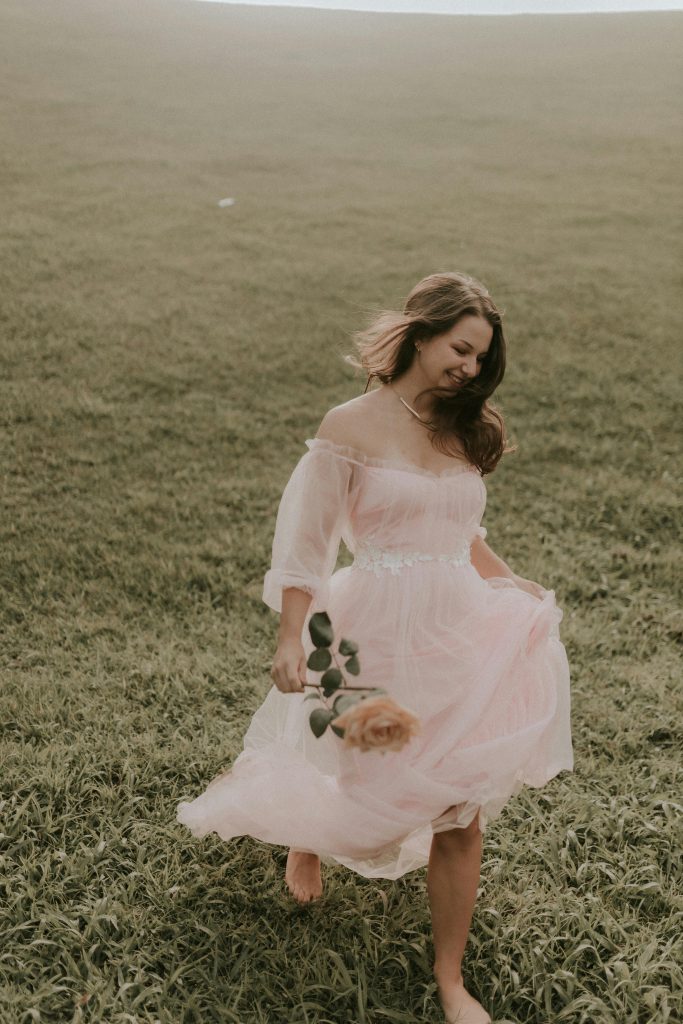 A young woman in a flowing pink dress walking barefoot in a grassy meadow holding a rose, enjoying the sunlight.