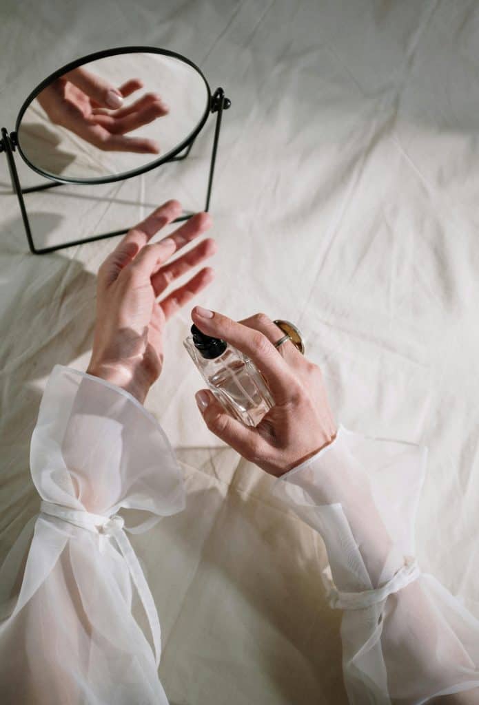 Hands in elegant sheer fabric holding perfume bottle near mirror, creating a graceful reflection.