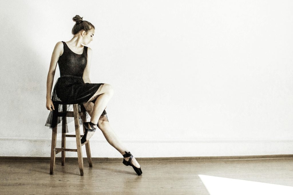 A ballerina in a stylish black dress sits elegantly on a stool in a minimalist studio setting.