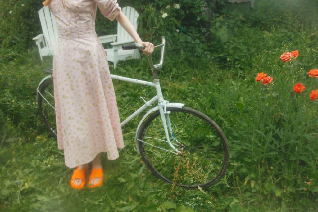 A woman in a floral dress holding a bicycle in a lush garden of flowers and chairs.